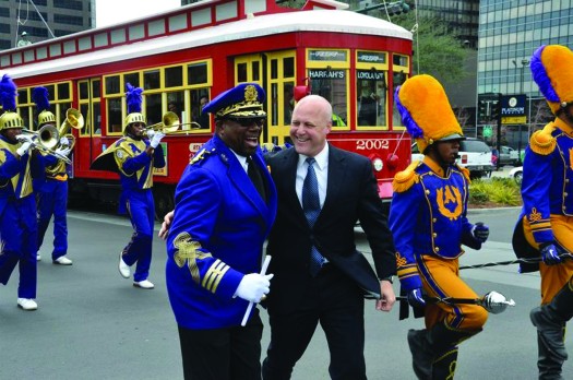 An image of Mitch Landrieu standing in the middle of a group of people who are playing various instruments. A streetcar is in the background.